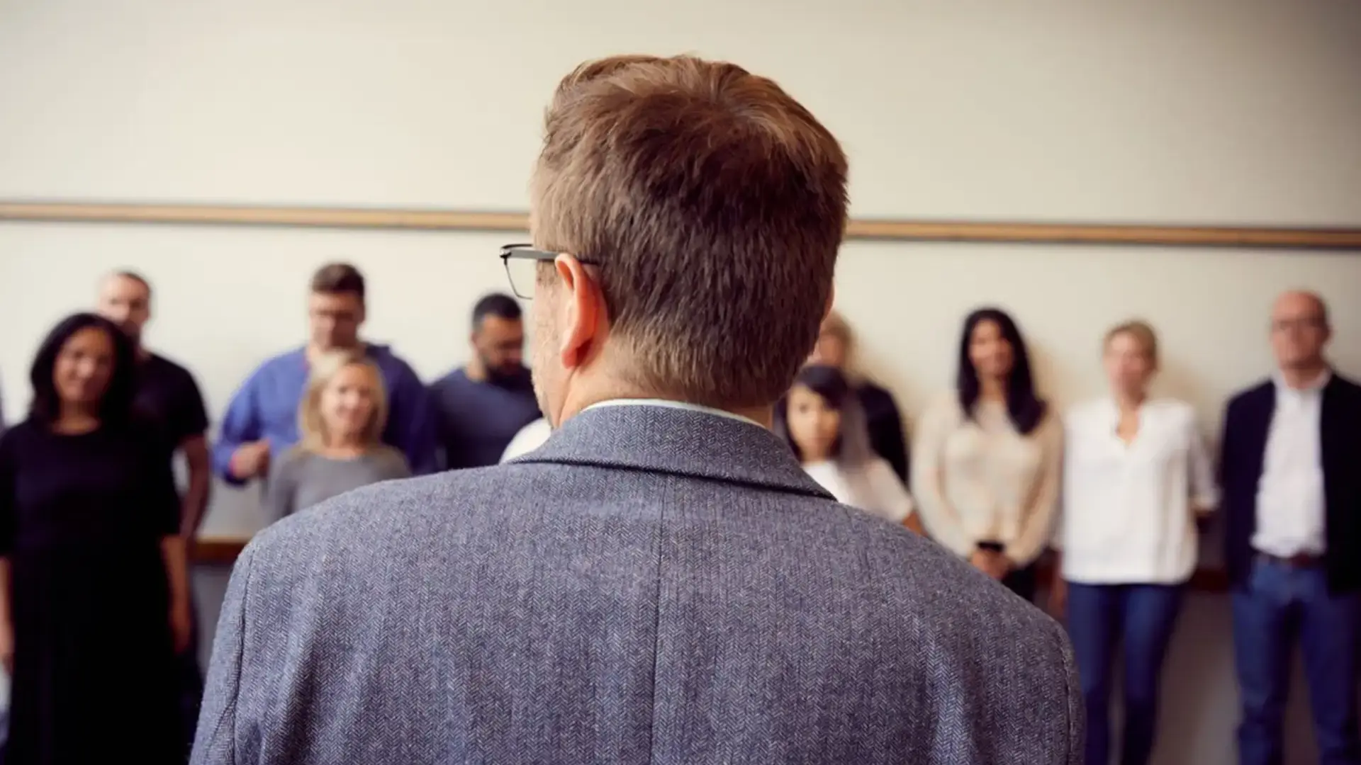 Man in a grey blazer, seen from behind, addressing a group of people standing along a wall.