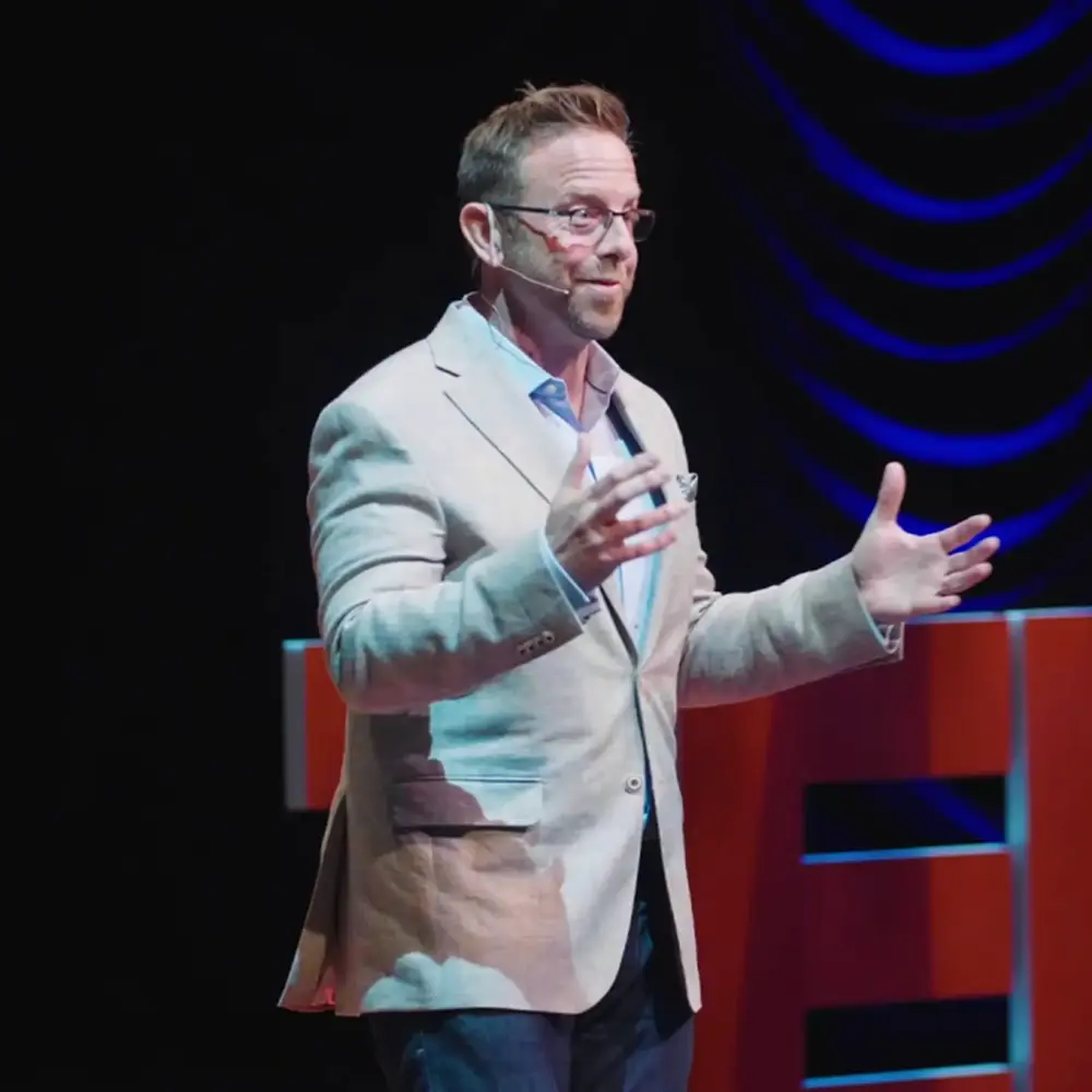 Speaker in a light blazer gesturing on a TED stage under blue lighting